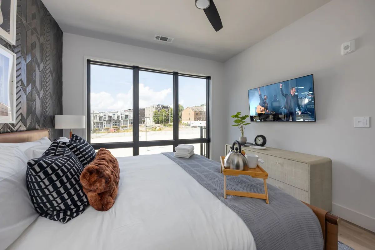 A contemporary bedroom with a king bed adorned with decorative pillows, large windows allowing in natural light, a wall-mounted smart TV, nightstand with lamp, and a decorative plant. The room exudes comfort and style, perfect for restful stays.