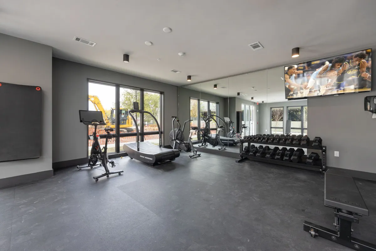 A bright, modern fitness center featuring treadmills, elliptical machines, a dumbbell rack, a TV, and mirrored walls. The area is well-lit, providing an inviting space for both individual workouts and group fitness sessions.