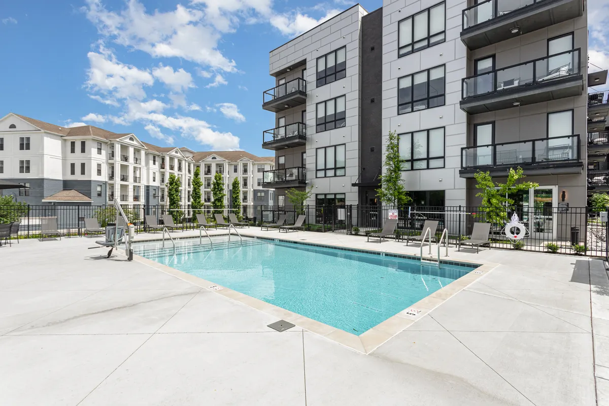 A sparkling swimming pool surrounded by lounge chairs and greenery, with balconies from the residential building overlooking the scene. Residents are seen swimming, sunbathing, and socializing by the pool, creating a vibrant, resort-like atmosphere.