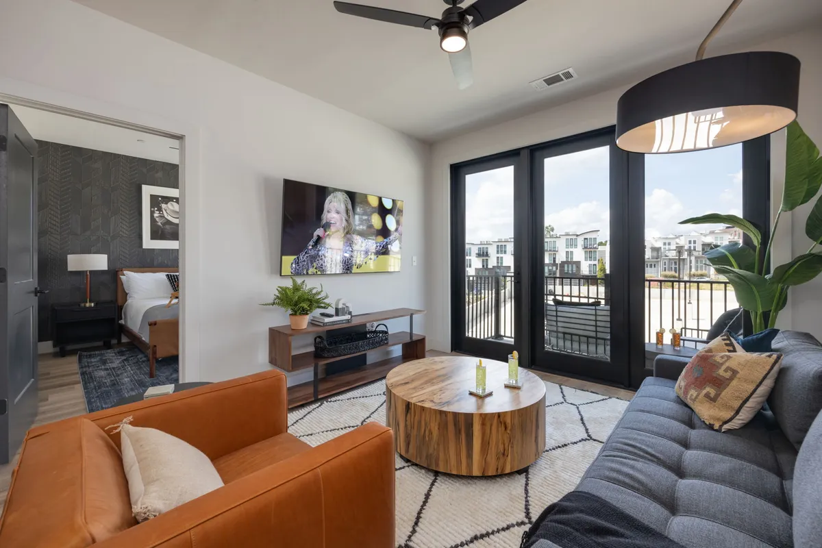 A cozy living room with large windows that allow abundant natural light to flow in. The space features a modern sofa, a coffee table, and a TV, creating an inviting setup for socializing, relaxing, or enjoying entertainment. The open arrangement hints at seamless transitions between living and adjacent spaces.