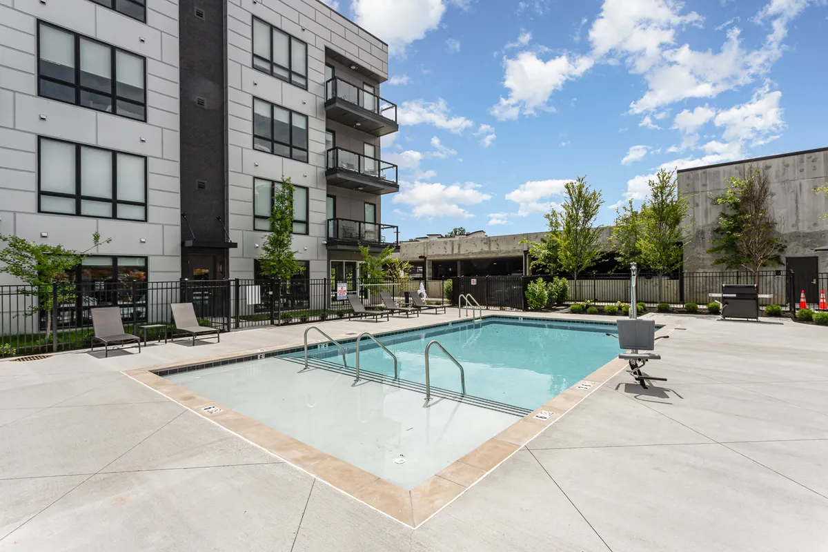 A wide view of the outdoor pool deck with lounge chairs, landscaped greenery, and the backdrop of modern apartment balconies. Residents are seen relaxing outdoors, suggesting a space designed for both activity and unwinding.