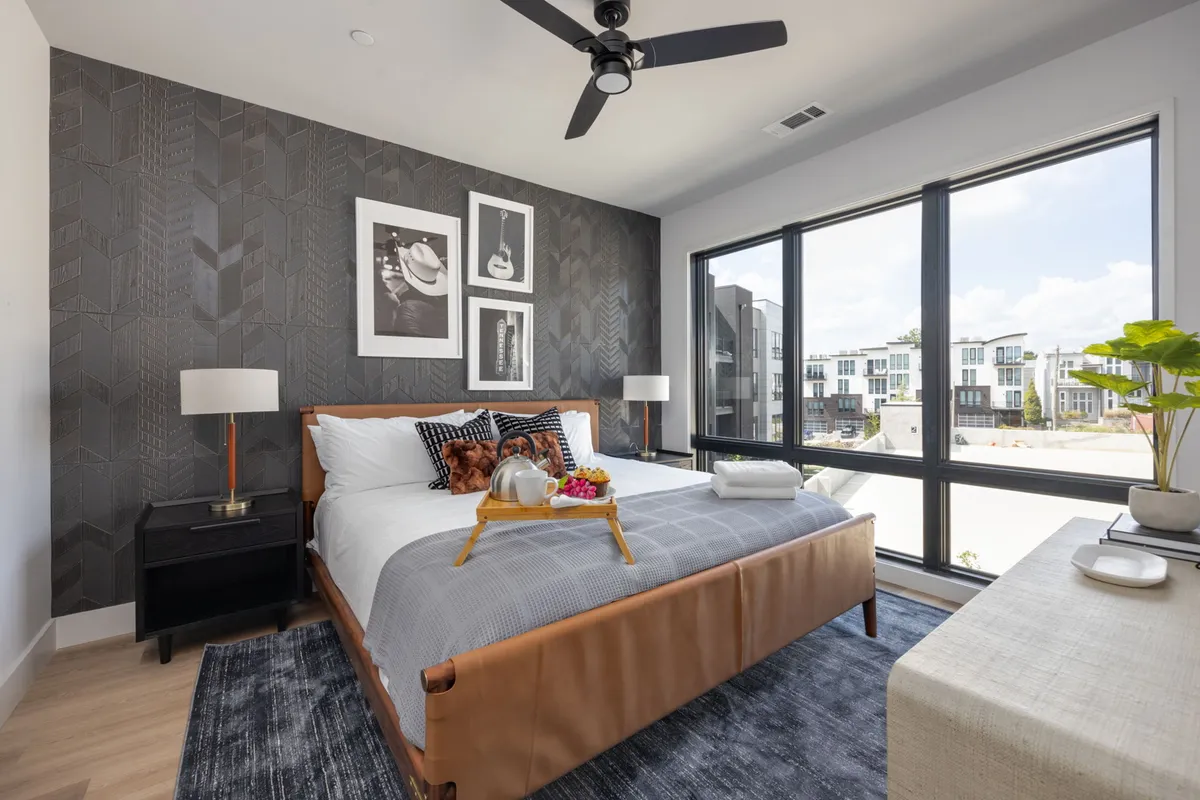 A cozy second bedroom featuring a plush queen bed, a window with city views, stylish artwork, a decorative plant on the table, and a nightstand with a lamp. The setting feels modern and inviting, ideal for guest comfort and privacy.