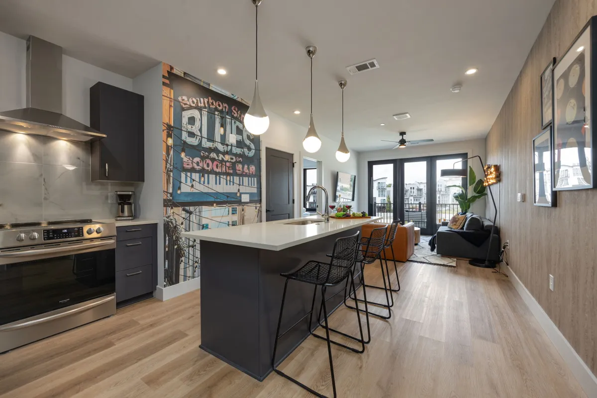 The image showcases a modern, open-concept kitchen and living space featuring a sleek island bar, vibrant wall mural, inviting seating area, and contemporary kitchen appliances. The space is flooded with natural light, suggesting a bright and lively atmosphere perfect for socializing and relaxation.
